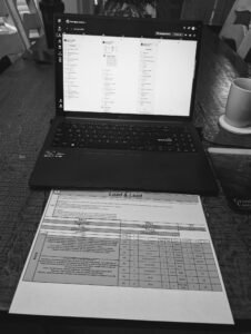 A close-up view of a laptop displaying a digital workout calendar next to a printed spreadsheet titled "Load & Lead." The spreadsheet shows a detailed 8-week cycle with percentages, sets, reps, and progression notes.