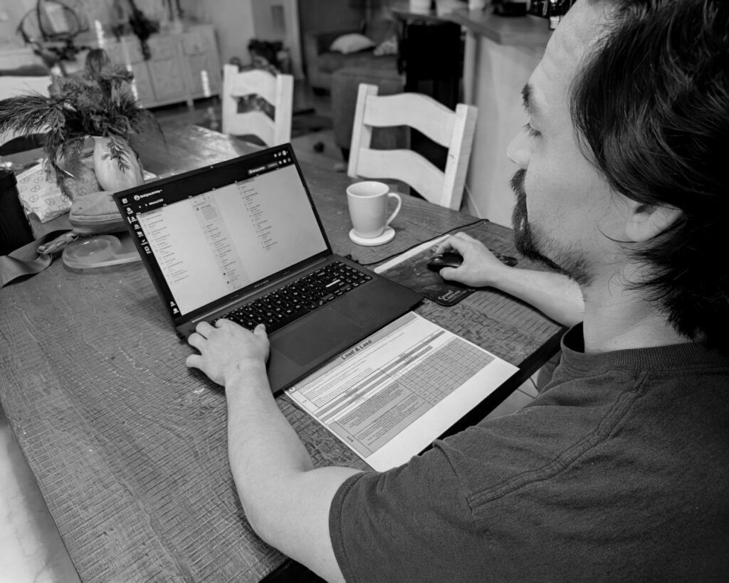 A black and white photo of a personal trainer sitting at a wooden table, focused on a laptop screen while referencing a printed program template titled "Load & Lead."