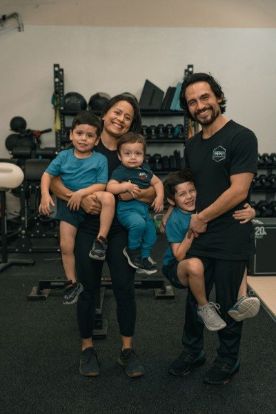A smiling family of five, including founders Austin Rodriguez and his wife, stand together in their new gym, Fitness Education & Training Center (FETC). Austin is holding one of their three young sons, while his wife holds the other two. They are surrounded by professional strength training equipment like dumbbells and squat racks in their Cedar Park facility.